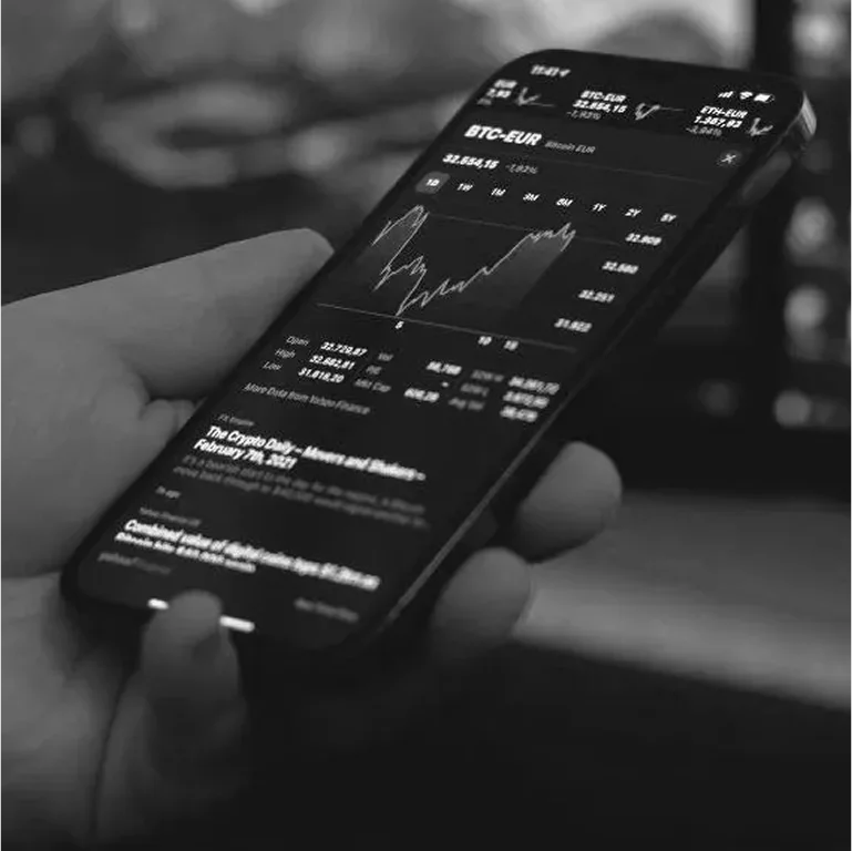 A person holding a smartphone displaying stock market charts and data in black and white.